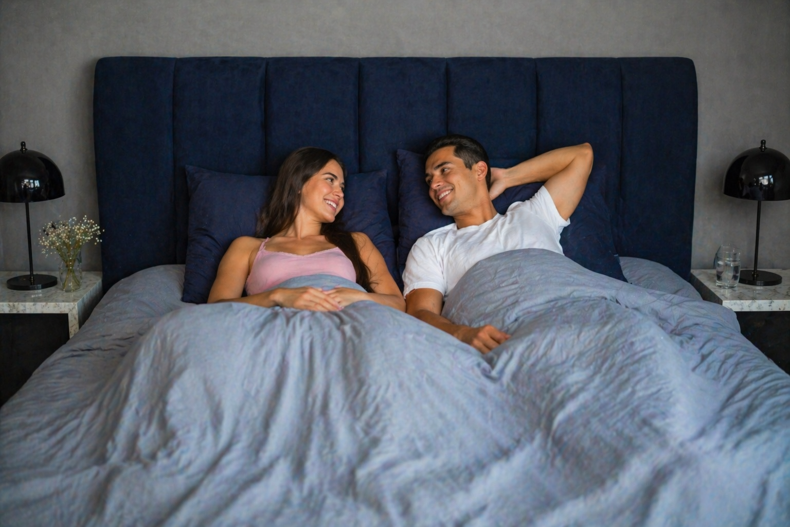 Glückliches Paar liegt morgens ausgeschlafen im Bett mit grauem Bettbezug und lächelt sich entspannt an in modernem Schlafzimmer