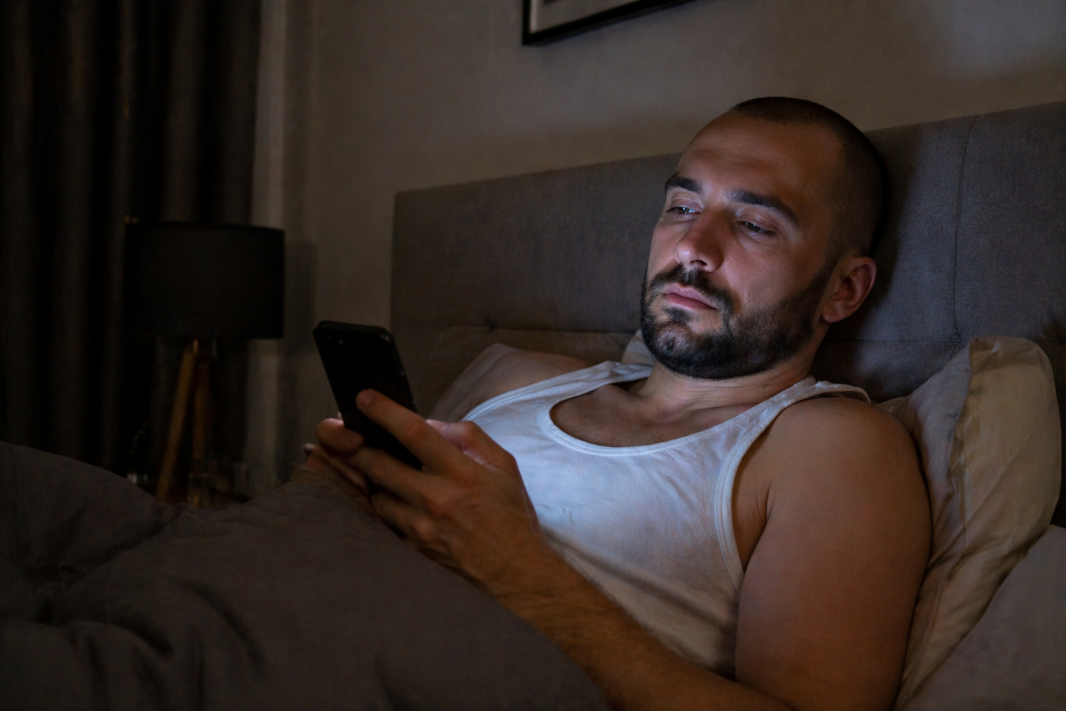 Müder Mann liegt im Bett und schaut auf sein Smartphone, dunkles Schlafzimmer mit Bildschirmlicht im Gesicht