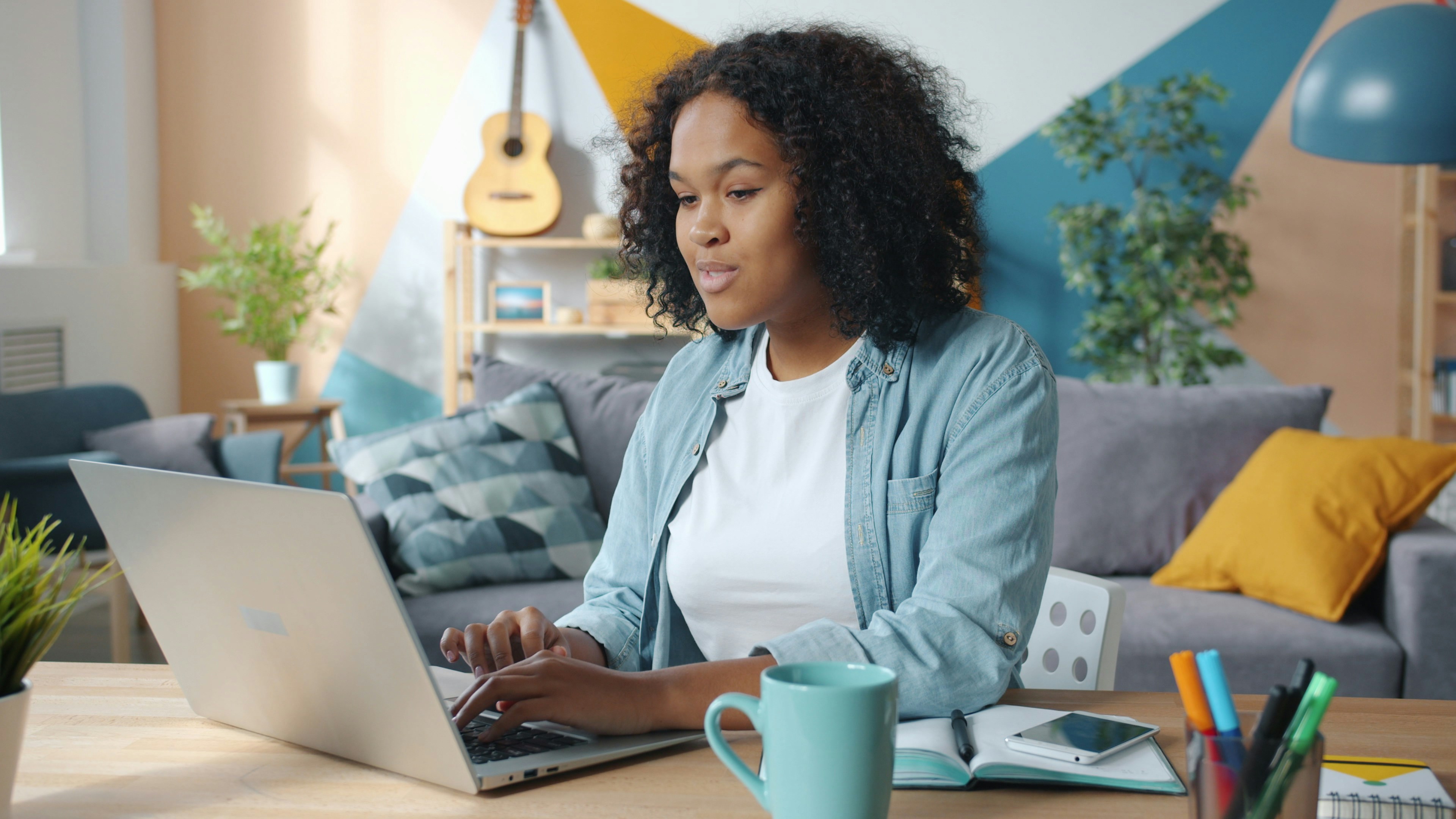 Eine Frau sitzt an einem Tisch im Wohnzimmer und arbeitet am Laptop. Vor ihr liegen ein Notizbuch, Stifte und ein Smartphone, daneben steht eine Tasse; im Hintergrund sind Sofa, Pflanzen und eine Gitarre zu sehen.