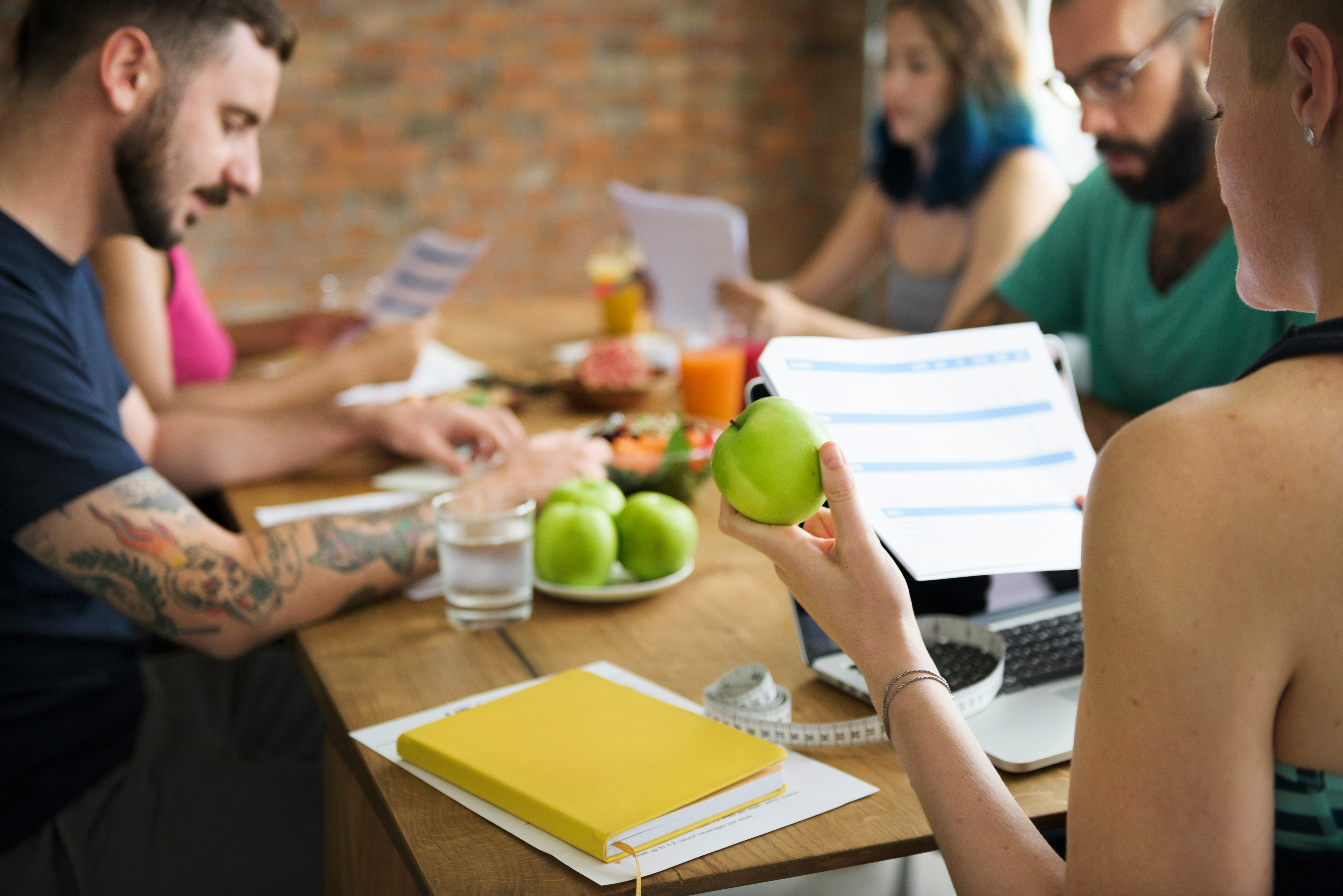 Learn@Home - Mehrere Erwachsene sitzen an einem Tisch und besprechen Unterlagen; im Vordergrund hält eine Person einen grünen Apfel neben einem Blatt mit Plan, auf dem Tisch liegen weitere Äpfel, ein Maßband, Getränke und ein gelbes Notizbuch.