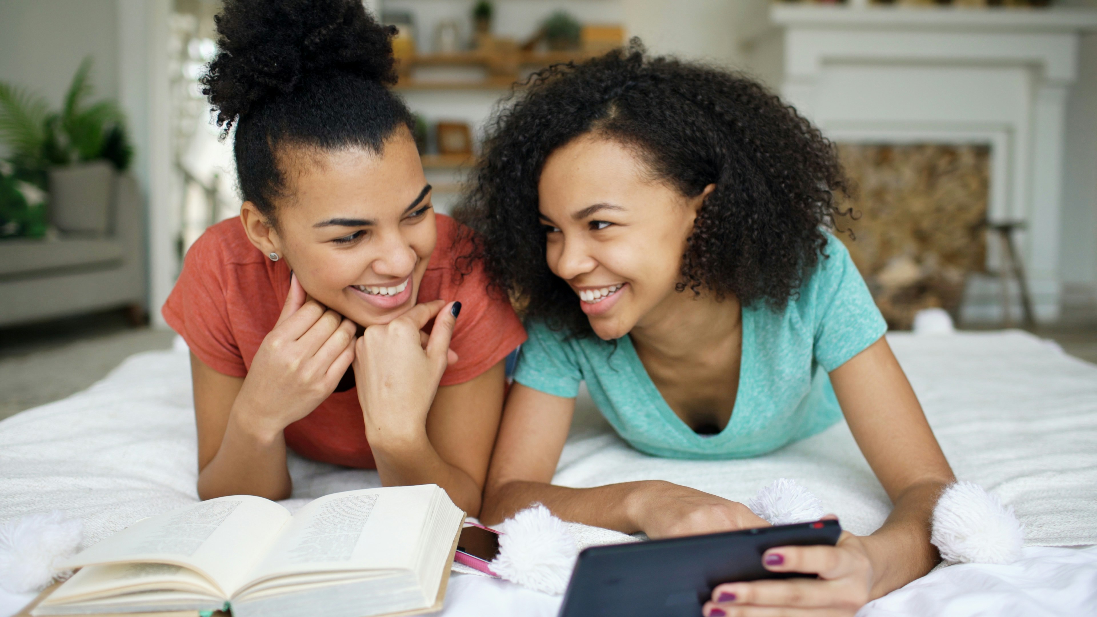 Learn@Home - Zwei junge Frauen liegen auf dem Bett, lachen und schauen auf ein Tablet; daneben liegt ein aufgeschlagenes Buch. Im Hintergrund ein helles Wohnzimmer.