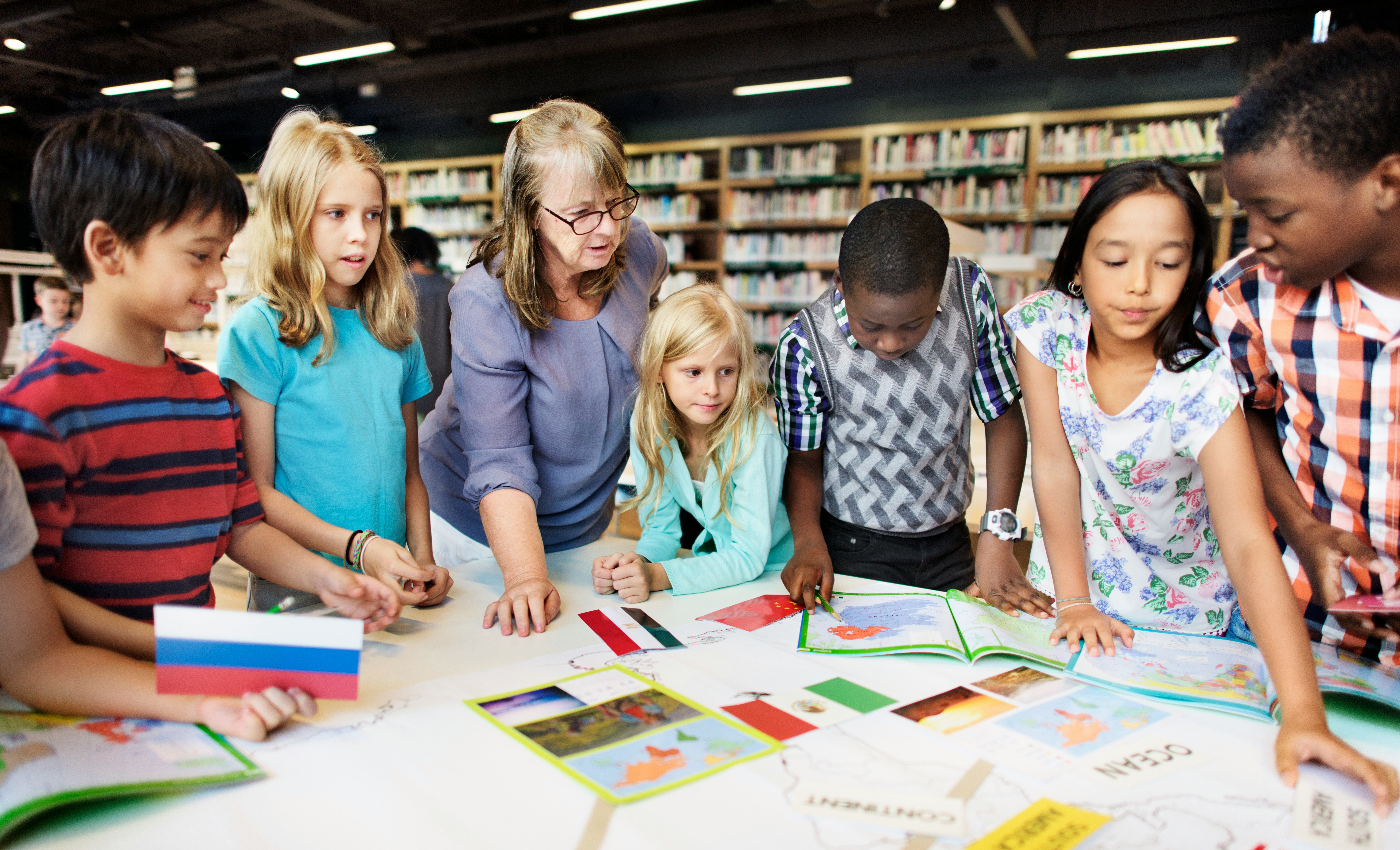Eine Lehrperson begleitet mehrere Kinder an einem Tisch in einer Bibliothek, während sie gemeinsam Karten und Materialien ansehen und an einer Aufgabe arbeiten.