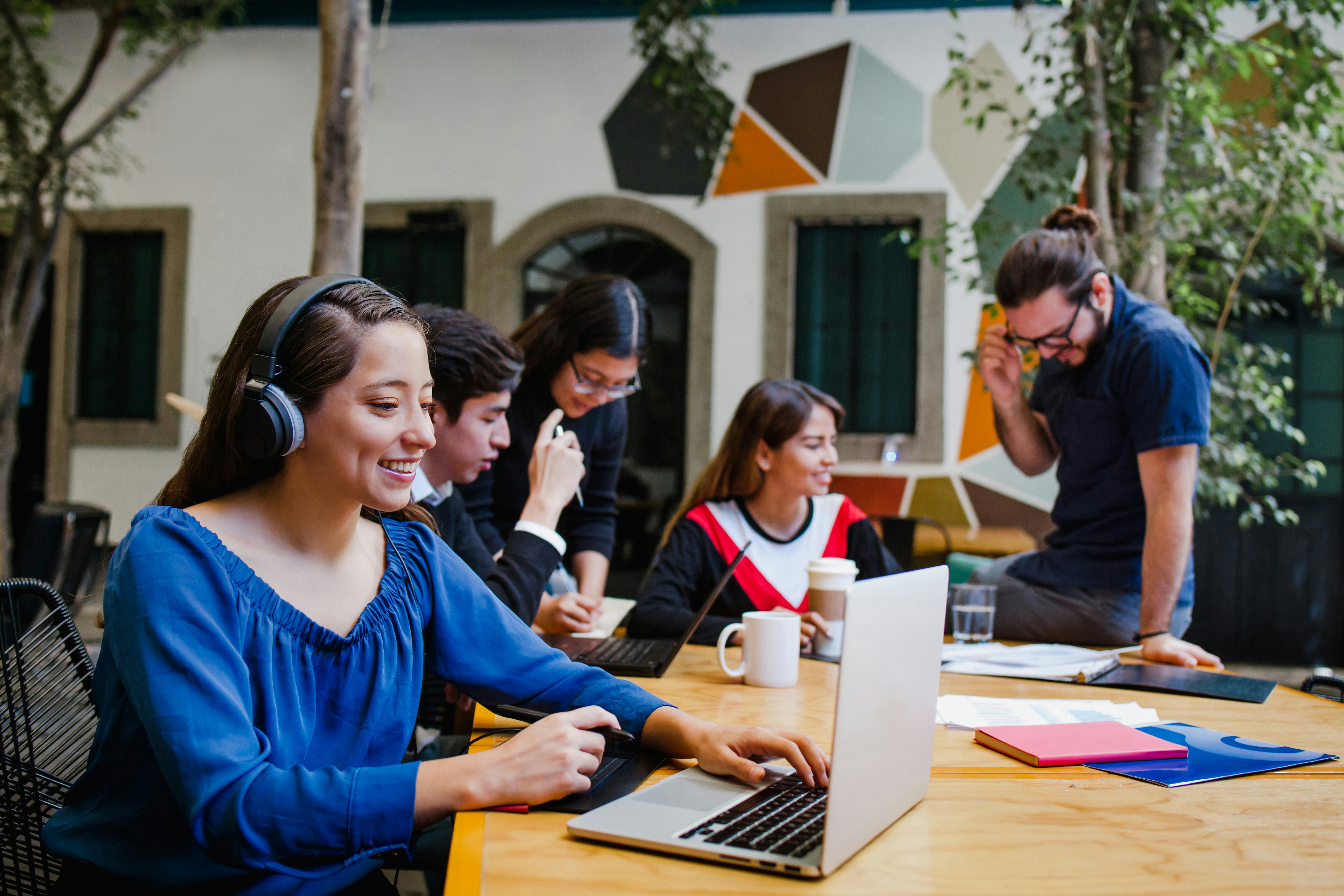 Learn@Home - Junge Frau mit Kopfhörern arbeitet am Laptop; im Hintergrund bespricht eine Gruppe am Tisch Unterlagen und Laptops. Szene in einem Innenhof mit Bäumen.