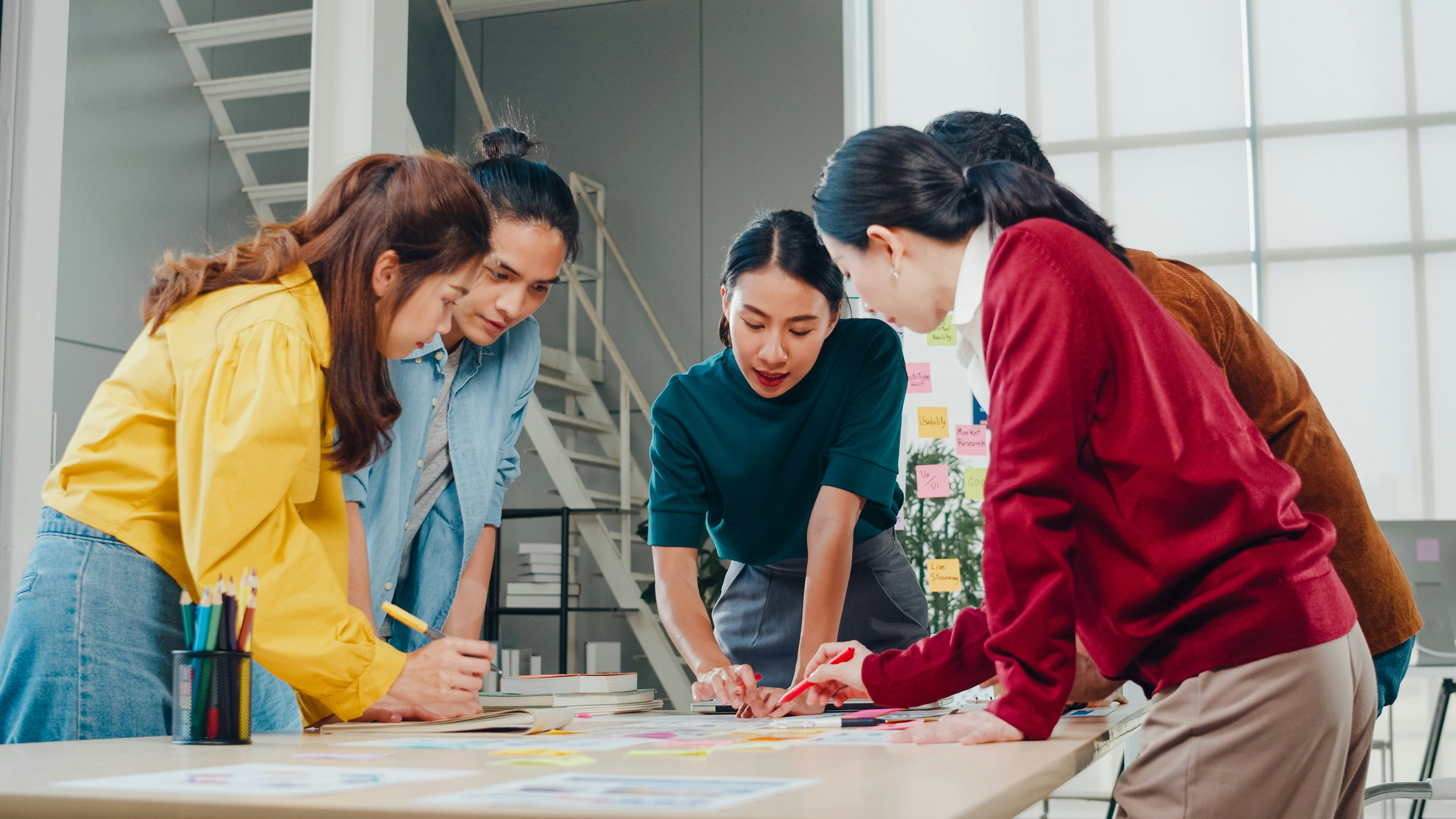 Learn@Home - Vier Personen stehen um einen Tisch und arbeiten an Notizen und Skizzen mit Stiften; im Hintergrund Treppe und Wand mit Haftnotizen in einem hellen Büro.