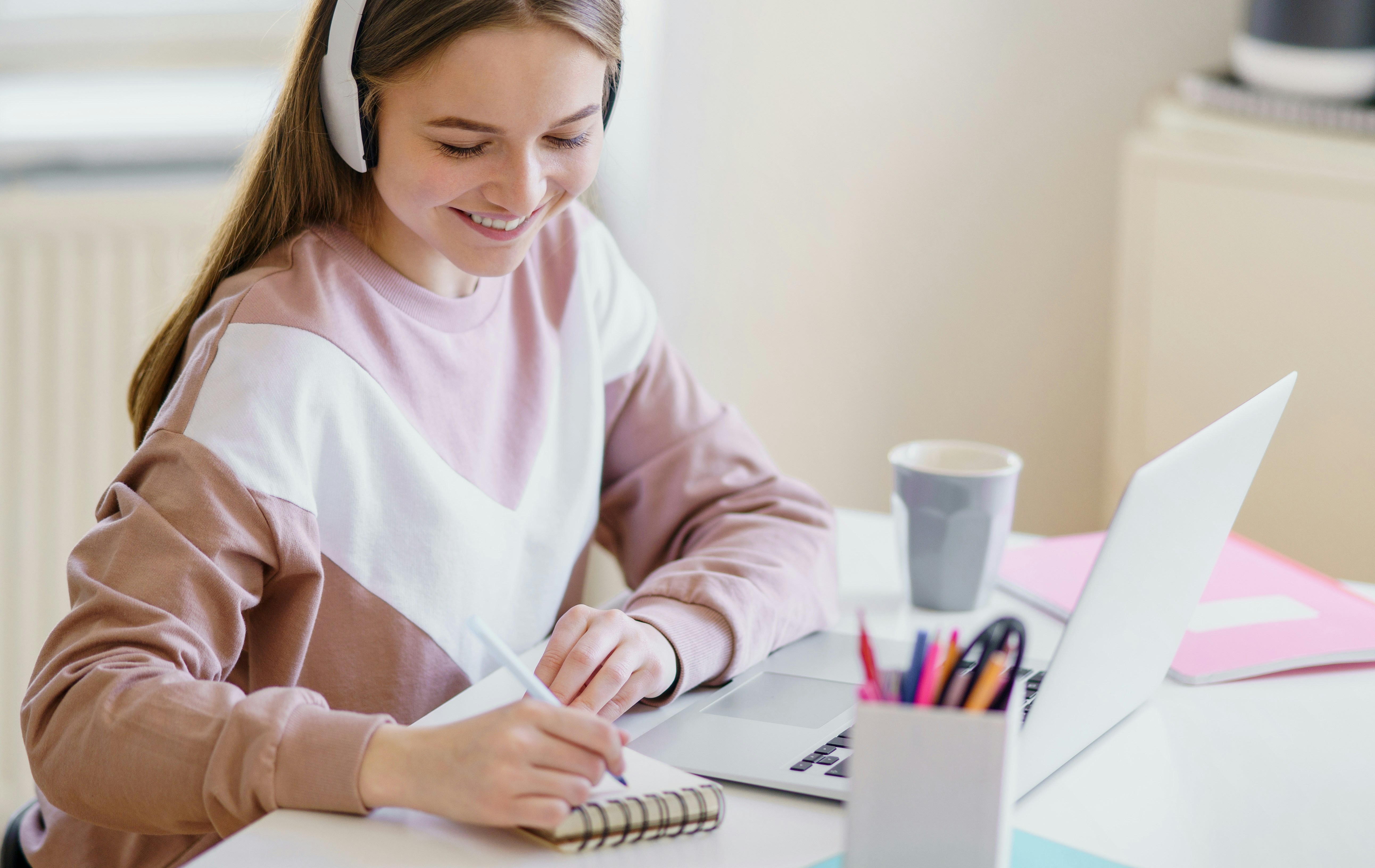 Learn@Home - Junge Frau mit Kopfhörern lernt zu Hause am Laptop und macht Notizen.