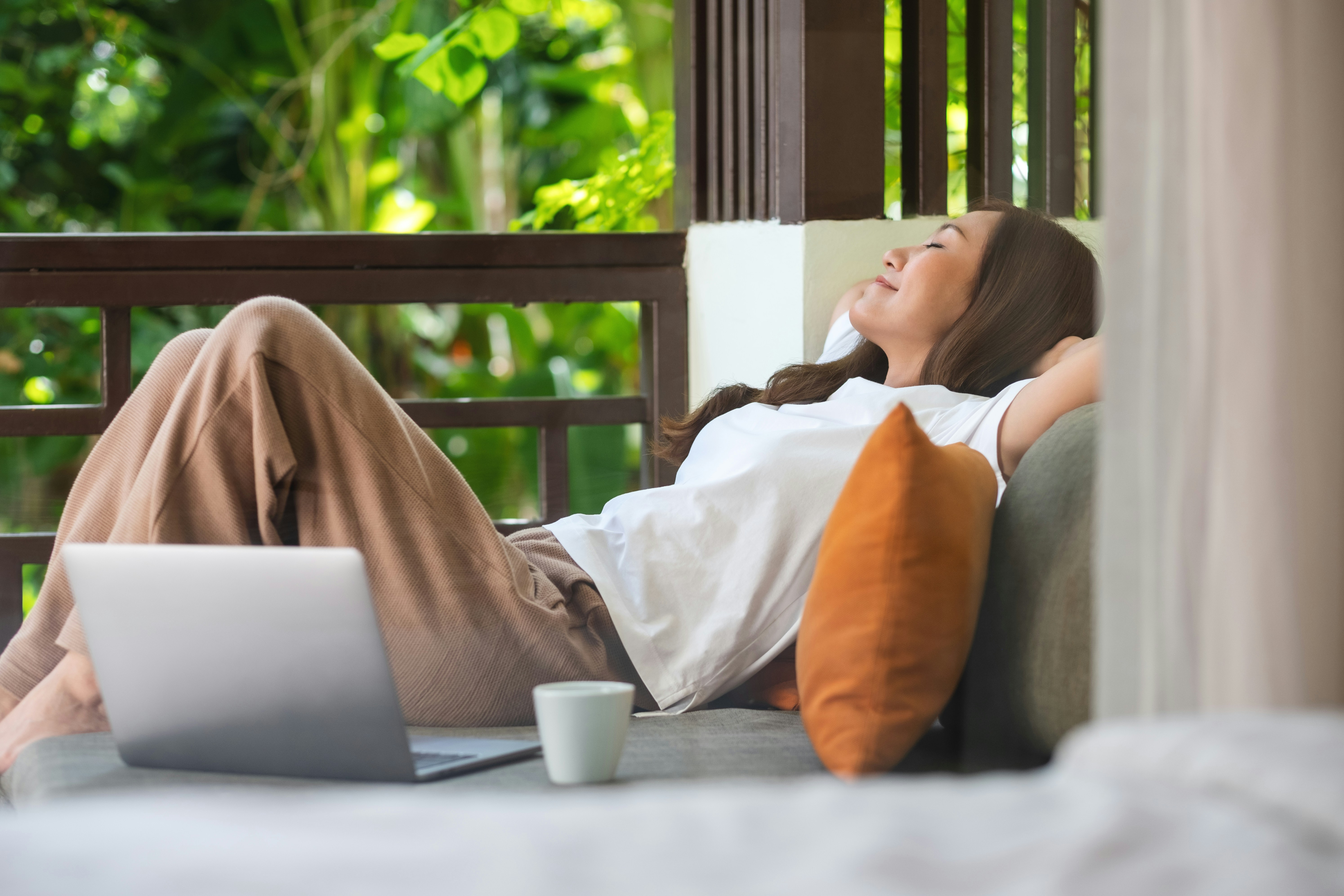 Learn@Home - Entspannte Frau mit geschlossenen Augen auf einer Couch neben Laptop und Tasse.