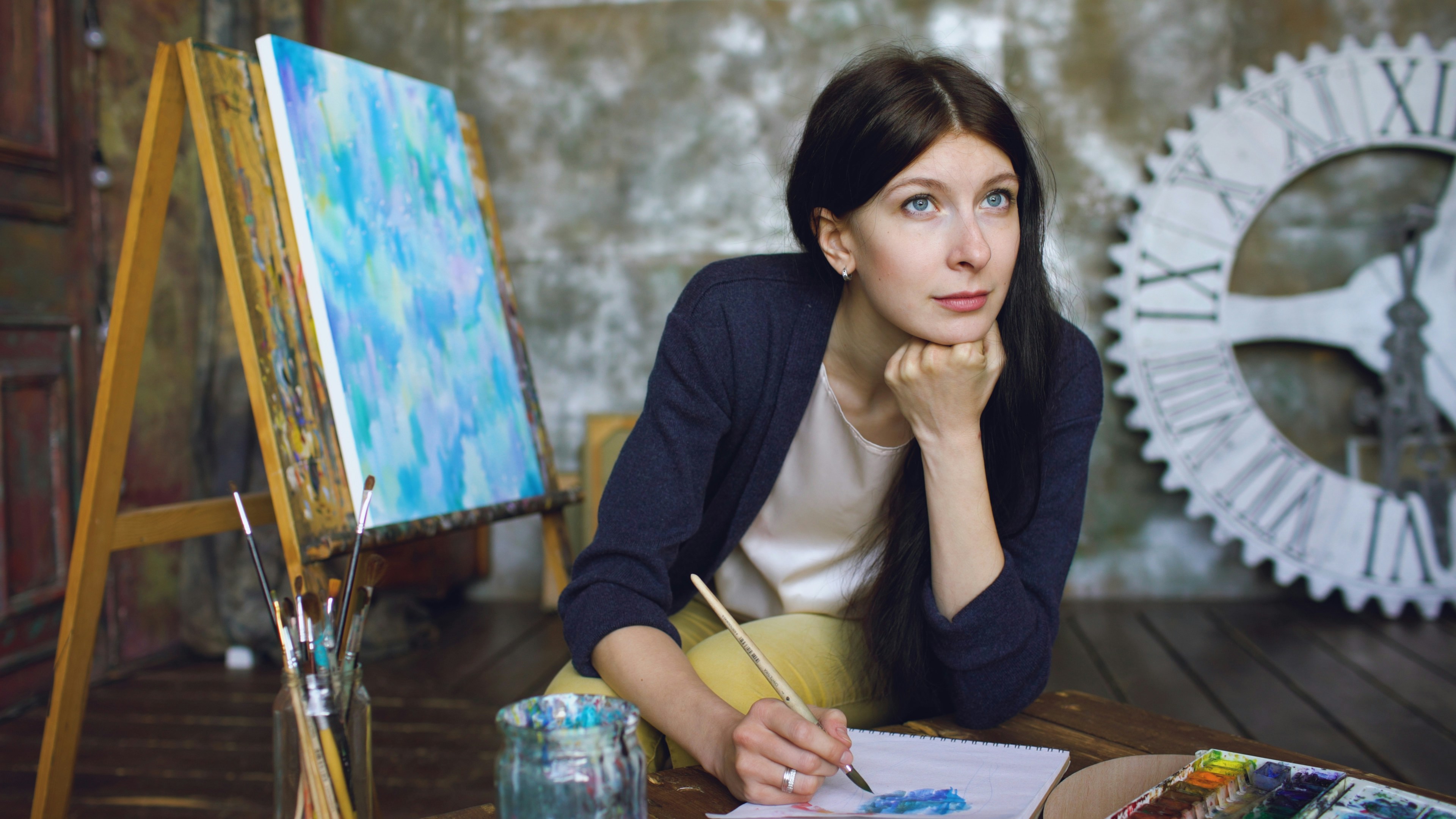 Learn@Home - Frau sitzt vor einer Staffelei und arbeitet mit Pinsel und Farben an einem kreativen Kunstprojekt.