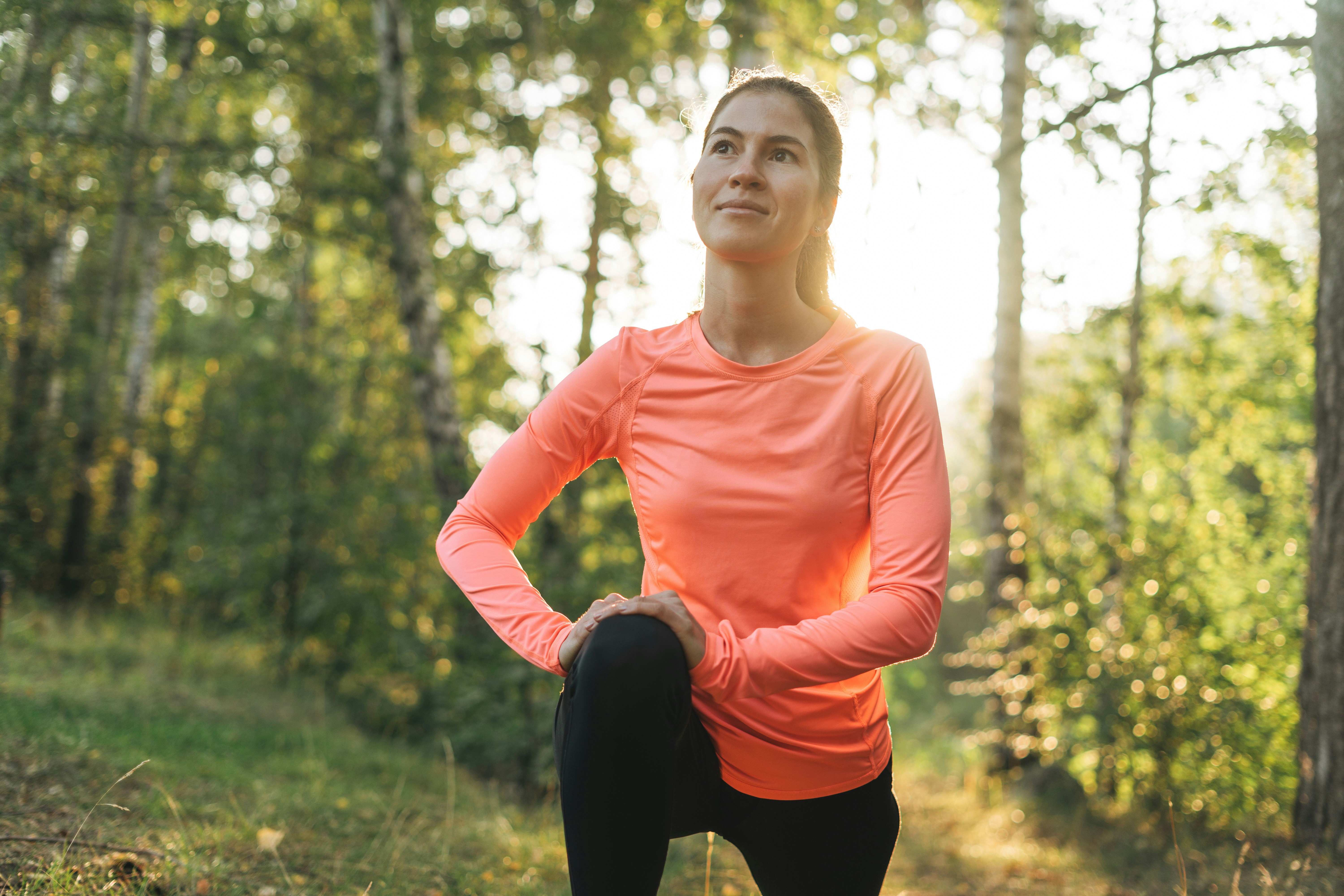 Learn@Home - Sportliche Frau in orangefarbenem Oberteil dehnt ihre Beinmuskulatur während eines Outdoor-Trainings in einem sonnendurchfluteten Wald.